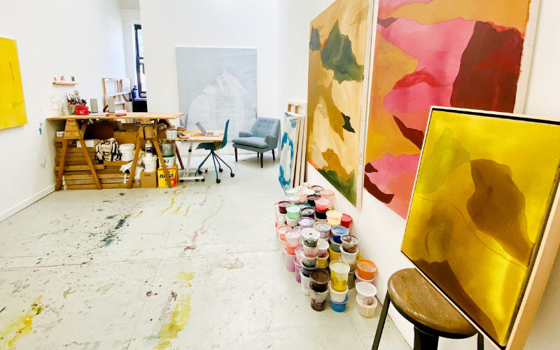 An inside view of artist Mel Reese's studio shows a vibrant, creative workspace. The studio features colorful abstract paintings leaning against the wall, with rich pinks, yellows, and greens. A wooden desk with art supplies, paint cans, and tools is set up on one side, alongside a chair and a lounge seat near a large, unfinished canvas. Numerous containers of paint are stacked on the floor