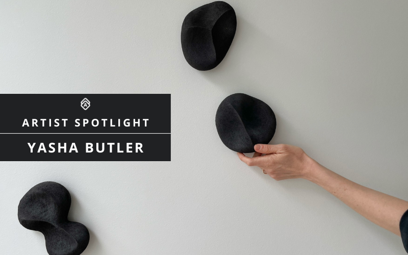 Three matte black ceramic sculptures mounted on a white wall, with a hand reaching to touch one piece. The sculptures appear organic and rounded in form. A black title card in the left of the image reads "ARTIST SPOTLIGHT" and "YASHA BUTLER" in white text, with a geometric logo.