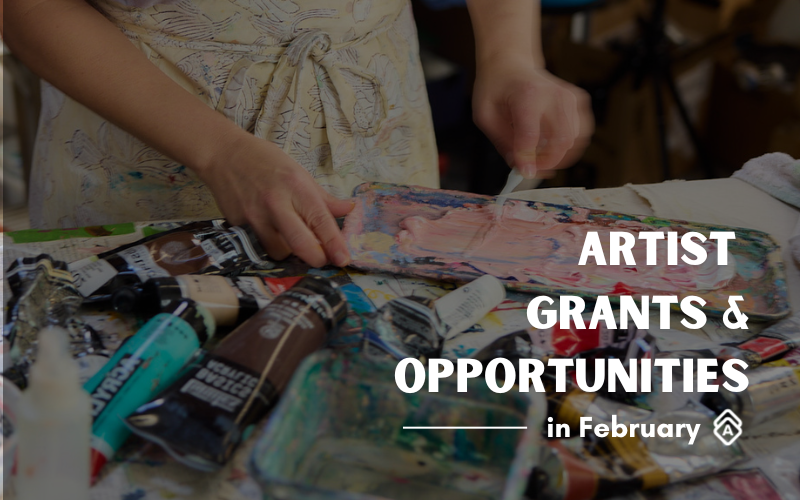 A close-up photo of an artist's hands working with paint tubes scattered on a surface. The artist wears a light-colored garment with pattern details. Paint tubes in various colors, including turquoise and brown, are visible in the foreground. Overlaid white text reads "ARTIST GRANTS & OPPORTUNITIES in February" with a small Artwork Archive logo below. The background is slightly blurred, creating focus on the hands and art supplies. Photo courtesy of Artwork Artist Mila Rossi.