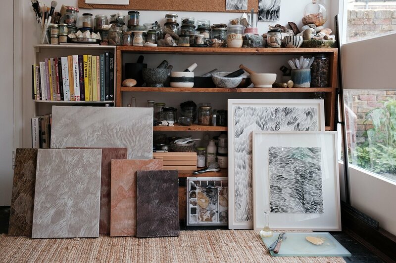 Lisa-Marie Price's artist studio features wooden shelving stocked with her creative materials. The top shelves house numerous glass jars containing pigments and natural materials, alongside her collection of mortars and pestles in black and white stone. Below are art books and several of her paintings leaning against the shelves, showing her characteristic textured patterns in muted tones of grey, brown, and pink. Framed artwork displaying similar feathered patterns sits to the right. A window provides natural light, and a sisal rug covers the floor space.