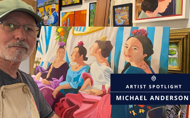 Artist Michael Anderson pictured in his studio wearing a paint-stained apron. He poses next to one of his colorful paintings, which depicts four women dressed in vibrant flamenco-style dresses with floral headpieces, seated against a blue-and-white striped backdrop. Behind him, framed paintings in a similar bright and expressive style fill the walls of his studio. A blue banner in the lower right corner reads 'Artist Spotlight: Michael Anderson.