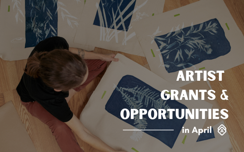 An overhead view of an artist's workspace promoting "Artist Grants & Opportunities in April." A person wearing a black top and burgundy pants is shown from above, sitting on a wooden floor surrounded by what appears to be cyanotype prints. The prints feature delicate botanical designs in white against deep blue backgrounds, with fern and leaf patterns clearly visible. Small green pieces of tape secure the prints to the floor. The text is overlaid in white capital letters on the bottom right of the image, with a decorative logo underneath. The composition suggests an artistic, DIY workspace where printmaking or alternative photographic processes are being explored.