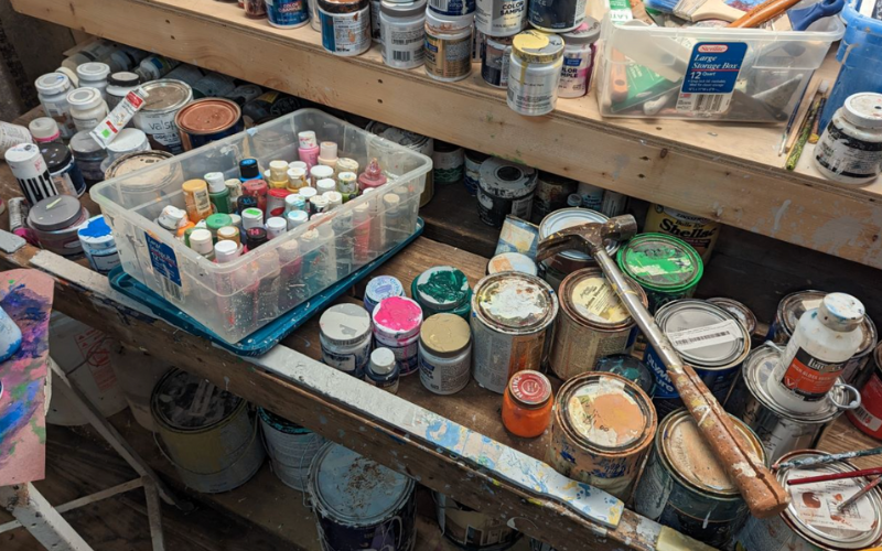 A wooden workbench covered with various cans, bottles, and jars of paint, including small acrylic paint bottles in a clear plastic container, larger paint cans with dried paint on their lids, and an assortment of color samples. A worn hammer with a paint-splattered handle rests on top of the paint cans. The workspace appears well-used, with splattered paint and supplies scattered around. 