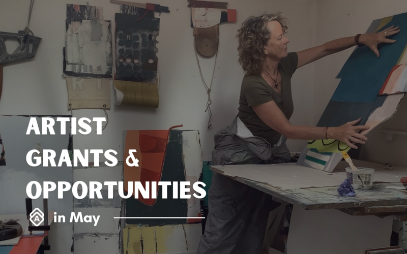 Text reads, "Artist Grants & Opportunities in May" featuring artist Karen Stamper at work in her studio. She has curly hair and wears an olive green t-shirt with gray pants, actively handling a blue artwork or canvas. The studio space shows her mixed media aesthetic with several abstract collage pieces hanging on the white wall behind her. A paint-splattered work table with supplies is visible in the foreground. The text "ARTIST GRANTS & OPPORTUNITIES" appears in bold white letters on the left side of the image, with "in May" in smaller text below, accompanied by a decorative logo. The composition captures the authentic working environment of a professional artist.