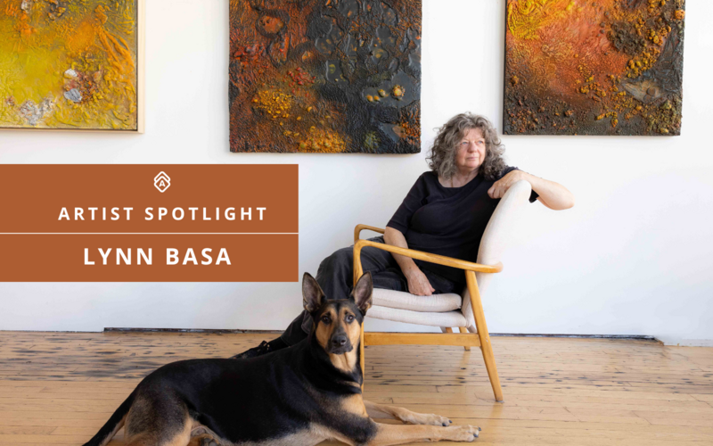 Artist Lynn Basa sits in a light wooden armchair in a bright studio with a polished wood floor, next to a large German Shepherd mix lying at her feet. Behind her are three textured, abstract mixed-media artworks in earthy tones of yellow, orange, and black. A rust-colored banner on the left reads "ARTIST SPOTLIGHT – LYNN BASA."