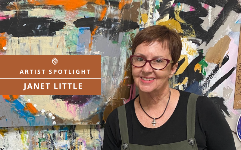 Artist Janet Little stands smiling in front of one of her bold, abstract acrylic paintings featuring layered textures and expressive brushstrokes in black, orange, cream, and pastel tones. She wears glasses, a black top, and green overalls with a cross necklace. A banner on the image reads "Artist Spotlight: Janet Little" with the Artwork Archive logo.
