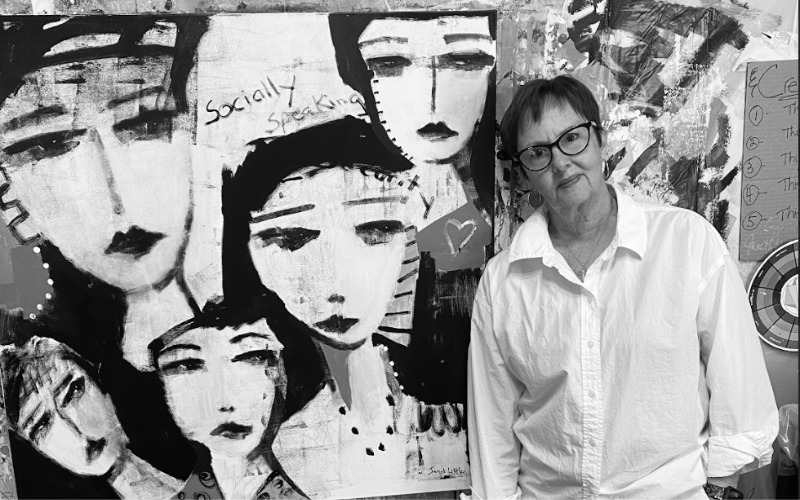 Black and white photo of artist Janet Little standing beside one of her expressive acrylic paintings. The artwork features five abstract female faces with bold, dark features and handwritten text that reads "Socially Speaking." Janet wears a white button-up shirt and glasses, standing in her studio surrounded by textured, layered canvases and a handwritten list tacked to the wall.