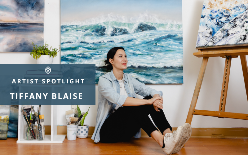 Artist Tiffany Blaise sits on the floor of her studio, smiling and looking off to the side. Behind her is a large, vibrant seascape painting featuring crashing ocean waves. She’s surrounded by art supplies, including brushes, paint containers, and other tools, with more artwork on the walls and an easel nearby. A text overlay reads: "Artist Spotlight – Tiffany Blaise.
