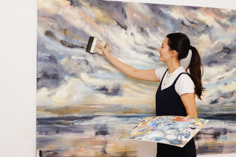 Artist Tiffany Blaise smiles while working in her studio, using a large paintbrush to apply dark brushstrokes to a dramatic, atmospheric landscape painting. She holds a palette filled with colorful mixed paints and stands in front of a canvas featuring swirling clouds and reflective water.