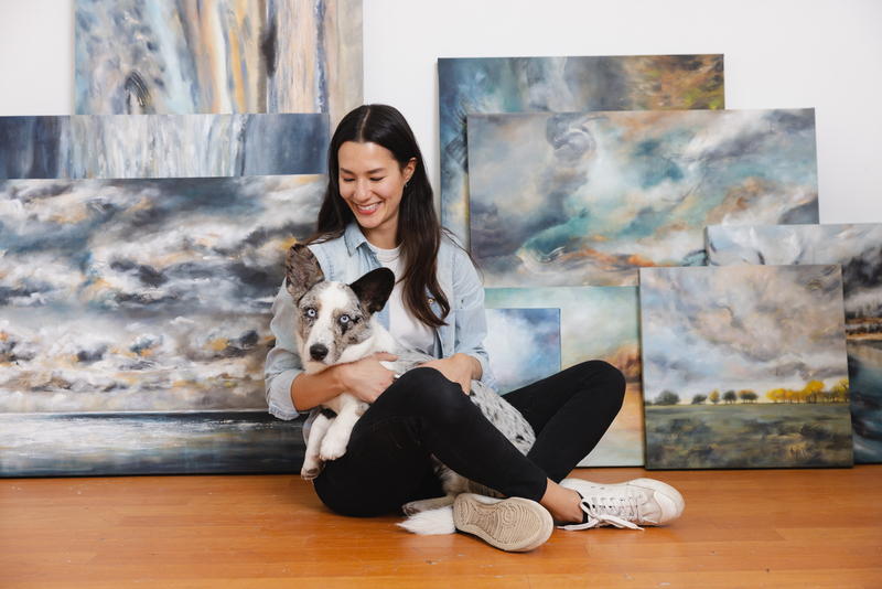 Artist Tiffany Blaise sits on the floor of her studio, smiling and holding her dog, a white and gray pup with striking blue eyes. Behind them is a display of her completed landscape paintings, featuring moody skies, abstract cloud forms, and natural scenes