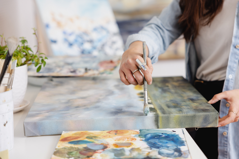 Close-up of artist Tiffany Blaise working on a landscape painting in progress. She holds a paintbrush in one hand and steadies the canvas with the other, adding detail to a cloudy sky and water scene. A colorful palette and a jar of brushes sit nearby on the white studio table.