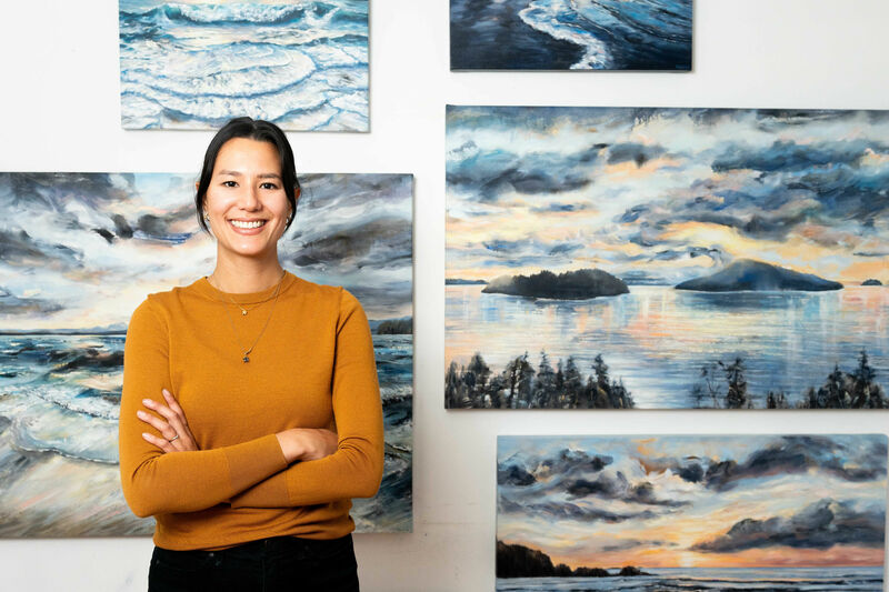 Artist Tiffany Blaise stands smiling with arms crossed in front of several finished landscape paintings featuring ocean scenes, moody skies, and coastal islands. She is wearing a mustard-colored long-sleeve shirt and black pants, and the background displays a vibrant collection of her expressive seascape and sky-themed artworks.