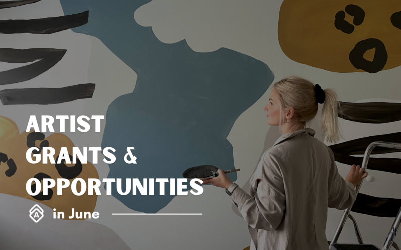 A banner graphic for "ARTIST GRANTS & OPPORTUNITIES in June." The text is prominently displayed in large white letters against a background featuring abstract painted shapes in blues, browns, and grays. On the right side of the image, a person with blonde hair in a ponytail is seen from behind/profile, wearing a light-colored jacket or smock. They appear to be painting on a large canvas or wall mural, holding what looks like a paint palette and standing on or near a step ladder. The composition suggests this is promotional material for upcoming arts funding or exhibition opportunities available to artists in the month of June, highlighting support for visual artists and creators.
