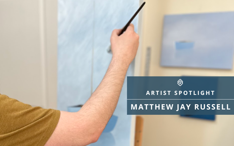  close-up image of artist Matthew Jay Russell’s arm as he paints a soft, minimalist seascape in shades of blue. His hand holds a thin paintbrush, mid-stroke, in front of a canvas on an easel. Another finished painting with a similar serene, blue-toned composition is visible in the background. Overlay text reads "Artist Spotlight: Matthew Jay Russell.