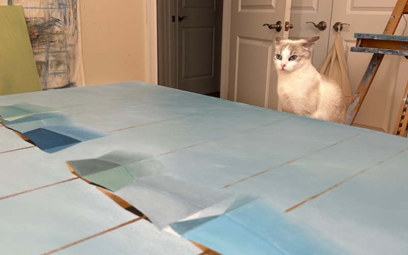 Inside artist Matthew Jay Russell’s studio, a curious white and gray cat with striking blue eyes—his “furry studio assistant”—sits alertly near a large artwork in progress. The painting, laid flat on a table, features a soft, geometric seascape in pastel blues and greens, with tape marking sections of the canvas.