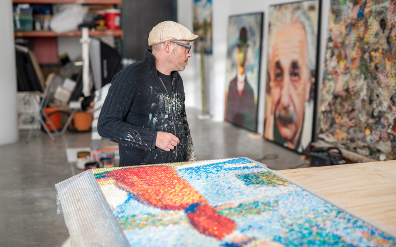 Artist Bradley Hart stands in a bright studio, wearing a paint-splattered sweater and cap, looking at a large, colorful artwork made from bubble wrap and acrylic paint. Several finished portraits lean against the wall in the background.