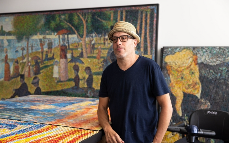 Artist Bradley Hart stands in his studio beside a table covered with a colorful bubble wrap artwork. Behind him are large-scale bubble wrap paintings, including a reinterpretation of Seurat’s pointillist park scene and another textured portrait. Hart wears a straw hat and dark t-shirt.