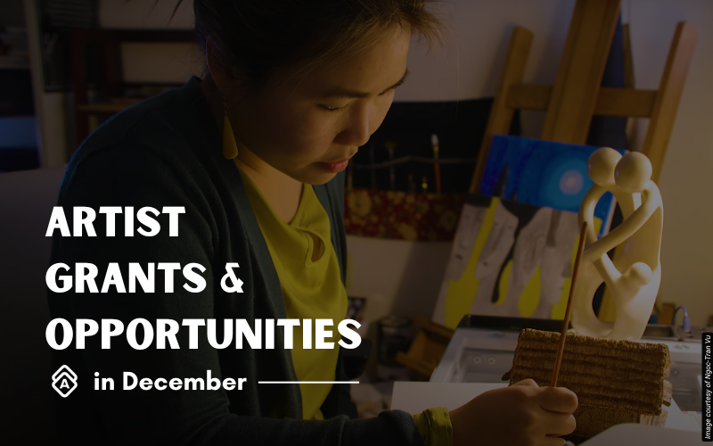 Artist Ngoc-Tran Vu works intently in her studio, wearing a bright yellow top. She holds what appears to be a textured material or artwork sample while examining it closely. The studio space contains wooden easels, colorful paintings, and art supplies scattered throughout. Behind her, canvases with blue and other vibrant colors are visible, along with various sculptural elements including what looks like white ceramic or plaster forms. The lighting creates a warm, focused atmosphere in the creative workspace. Text overlay reads "ARTIST GRANTS & OPPORTUNITIES in December" in white letters against the left side of the image.