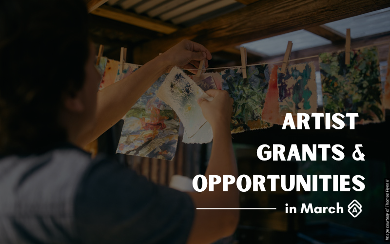 Artist Thomas Flynn II works in his studio, hanging colorful artwork on a clothesline with wooden clothespins. He wears a dark shirt and is shown from behind as he arranges what appears to be painted or printed paper pieces. The studio has exposed wooden ceiling beams and natural lighting from skylights above. Multiple artworks featuring vibrant greens, blues, oranges, and other colors are clipped to the line, displaying various abstract patterns and textures. The workspace has a rustic, intimate feel with its traditional drying method for displaying works. Text overlay reads "ARTIST GRANTS & OPPORTUNITIES in March" in white letters on the right side of the image.