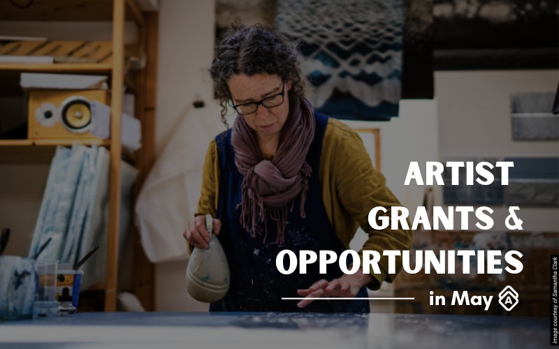 Artist Samantha Clark works in her studio, wearing glasses, a mustard yellow cardigan, and a purple scarf. She has curly dark hair and is focused on handling what appears to be a ceramic vessel or sculptural piece. The studio contains various artworks including geometric black and white patterns, blue and light-colored pieces, and other mixed media works displayed on walls and surfaces. Wooden shelving and storage areas are visible in the background, creating an organized creative workspace. The artist appears engaged in examining or working with three-dimensional objects. Text overlay reads "ARTIST GRANTS & OPPORTUNITIES in May" in white letters on the right side of the image.