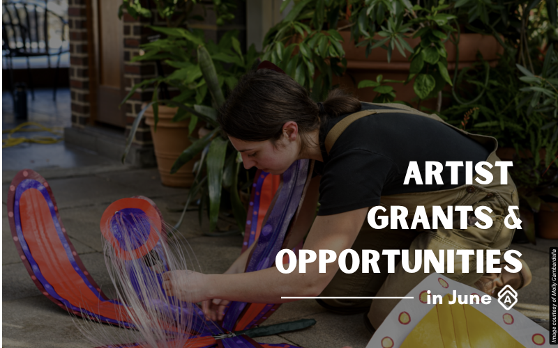Artist Molly Gambardella works outdoors in a courtyard setting, kneeling beside a large sculptural piece with flowing orange and purple ribbons or fabric elements. She wears a black top and has long dark hair as she carefully arranges or adjusts the colorful installation. The urban environment features brick buildings, lush green plants in terracotta pots, and concrete flooring. The artwork appears to be a three-dimensional piece with vibrant streaming elements that create dynamic movement. The setting combines industrial architecture with abundant greenery, providing an inspiring outdoor workspace. Text overlay reads "ARTIST GRANTS & OPPORTUNITIES in June" in white letters on the right side of the image.