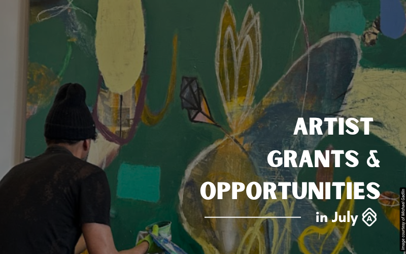 Artist Michael Gadlin works on a large-scale abstract painting in his studio, viewed from behind as he applies paint to the canvas. He wears a dark shirt and has dark hair while focusing on the artwork. The painting features bold organic forms in greens, yellows, and earth tones with flowing, botanical-inspired shapes and layered brushwork. The composition includes oval and leaf-like forms that create dynamic movement across the canvas surface. The studio setting appears spacious with good lighting for working on large contemporary paintings. Text overlay reads "ARTIST GRANTS & OPPORTUNITIES in July" in white letters on the right side of the image.