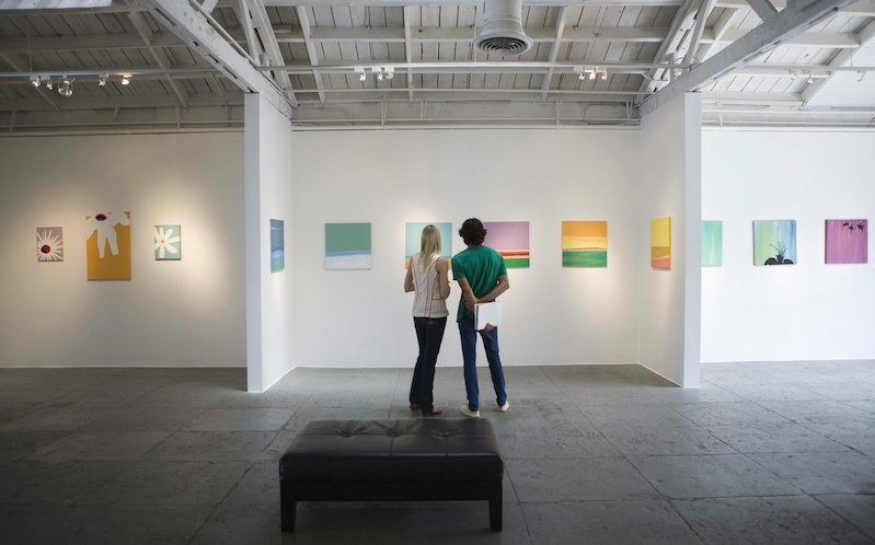 two people look at colorful paintings on a white gallery wall