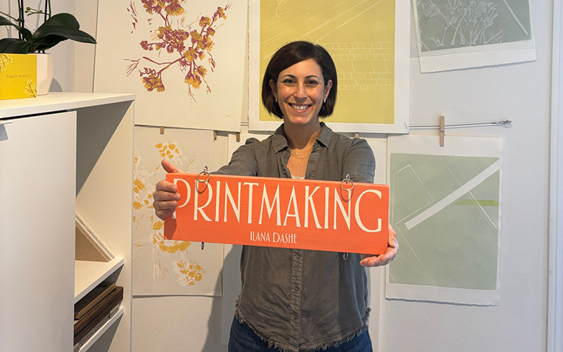 An artist in her studio holding up a sign that says Printmaking