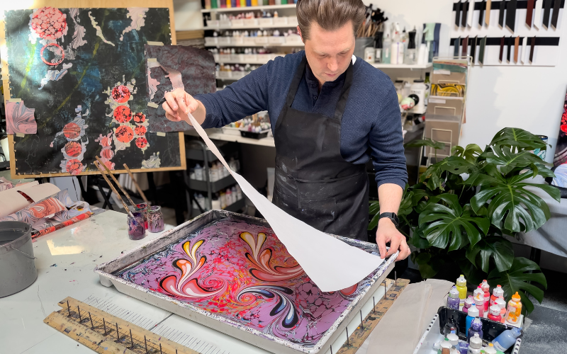 Featured Artist Keith Garubba lifts a freshly marbled sheet from the tray at Whistling Studios, revealing the moment the pattern transfers from water to paper. Photo courtesy of the artist.