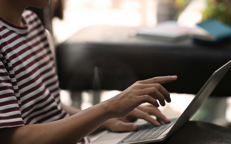 Cropped image of a person in a striped shirt with a laptop in their lap, and they are pointing towards the screen with one hand and have the other hand on the keyboard
