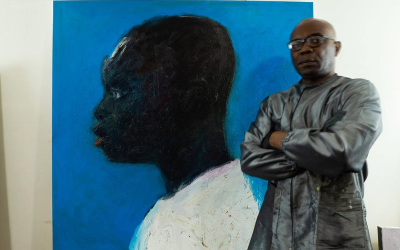Artist Temi Wynston Edun stands beside one of his oil stick paintings, a Black figure shown in profile against a deep blue background.
