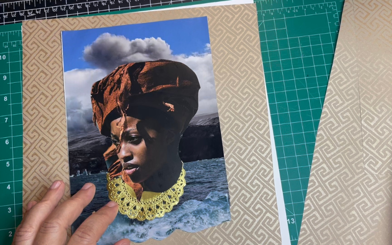 A WIP Gaskin collage rests on a studio cutting mat, showing a Black woman in a rust-toned headwrap and gold lace collar set against clouds and ocean.